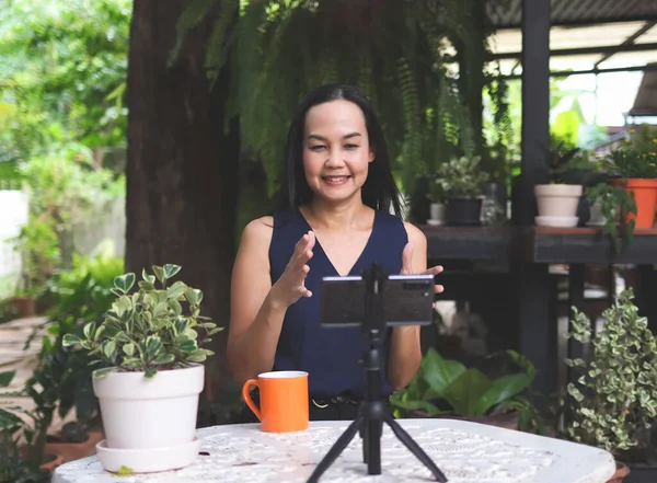 Portrait of Asian  woman coach or speaker, sitting at table in garen,  record live video broadcast on modern smartphone gadget, positive Asian woman  shoot webinar or online training course on cellphone device on tripod.