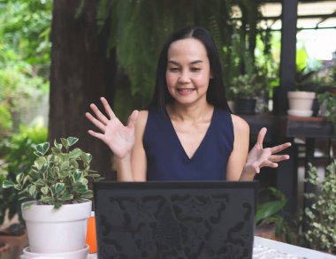 Bahçedeki masada oturan Asyalı kadının portresi, bilgisayar defteriyle video görüşmesi yapmak, bilgisayar monitöründe konuşmak ve işaret etmek..