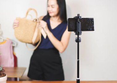 Evde çalışan Asyalı bir kadın, cep telefonuyla dokuma çantasını satıyor ya da video kaydediyor. Kapalı, seçici odaklanma. İş ve çevrimiçi satış kavramı.