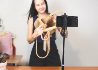 Evde çalışan Asyalı bir kadın, cep telefonuyla dokuma çantasını satıyor ya da video kaydediyor. Kapalı, seçici odaklanma. İş ve çevrimiçi satış kavramı.