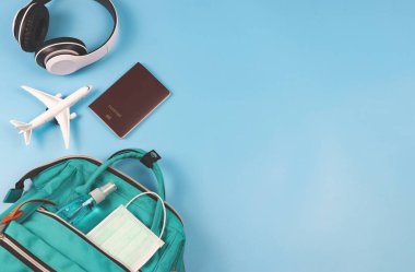 Üst ya da düz pasaport, kulaklık, uçak modeli ve tıbbi yüz maskesi ve mavi arka planda yeşil sırt çantalı alkol spreyi. Covid-19 salgını sırasında seyahat etmek.