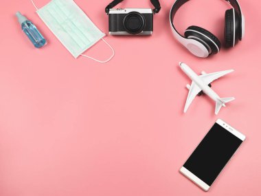 Uçak modeli, kamera, kulaklık, cep telefonu, koruyucu yüz maskesi ve pembe arka planda alkol spreyi. Yolculuk sırasında Covid19 koruması .