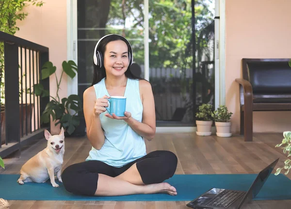 Portrait of Asian woman wearing headphones, holding coffee cup, sitting  on yoga mat in balcony  with computer laptop Chihuahua dog. Yoga exercise training online