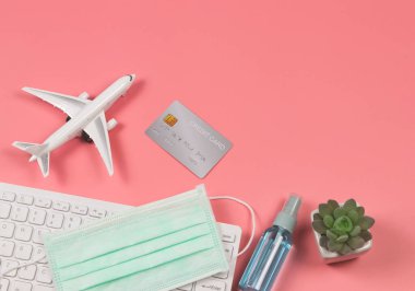 Üst ya da düz uçak modeli, kredi kartı, bilgisayar klavyesinde tıbbi yüz maskesi, alkol spreyi ve pembe arka planda kaktüs. Covid-19 sırasında seyahat.