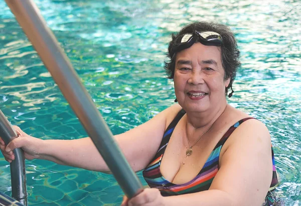 Portrait of happy and healthy Asian senior woman wearing colorful swimming wear and goggles, standing at stair, smiling and looking at camera. Elderly sports and active lifestyle.