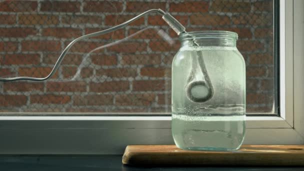 Processen för kokning av vatten i en glasburk genom uppvärmning av pannan från elektricitet, en stark ånga av bubblor, bakgrundstegel vägg och plastfönster, i förgrunden av plankorna — Stockvideo
