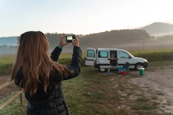 Young pretty woman taking a picture with her phone traveling by camper van though the countryside. Self built off-grid motorhome. Van life wild sunset.