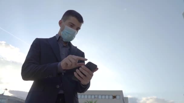 古典的なスーツでビジネスマンの閉鎖は屋外でスマートフォンを使用しています オフィスビルに立って携帯電話でビジネスコールを持っている コロナウイルスパンデミック対策顔マスク — ストック動画