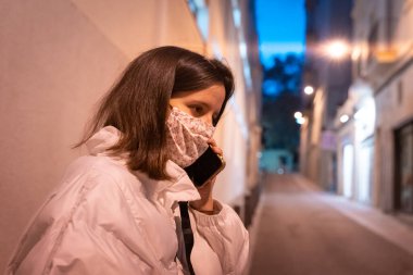 Gece şehir sokağında, ameliyat maskesiyle telefonunu kullanan ve kağıt bardakta kahve içen güzel genç bir kadın..