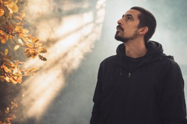 Yakışıklı genç adam sonbahar güneşli sabahında sisli ormanda tek başına yürüyor. Sıcak vintage renkler.