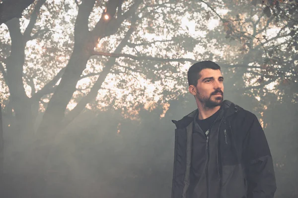 Yakışıklı genç adam sonbahar güneşli sabahında sisli ormanda tek başına yürüyor. Sıcak vintage renkler.