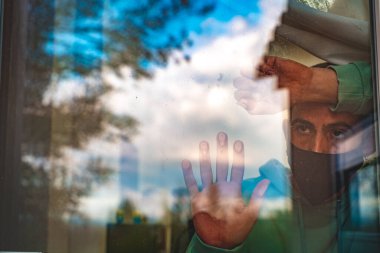 Coronavirus. Corona virüsünün hasta adamı pencereden bakıyor ve cerrahi maske takıyor ve hastalıktan kurtuluyor. Karantina hastası.