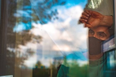 Coronavirus. Corona virüsünün hasta adamı pencereden bakıyor ve cerrahi maske takıyor ve hastalıktan kurtuluyor. Karantina hastası.