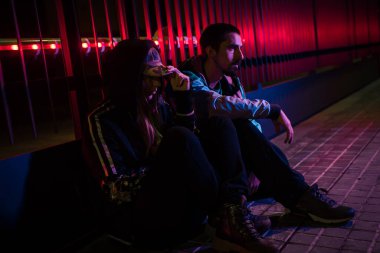 Neon ışıklı çitlerdeki şık genç bir çiftin güzel sinematik portresi. Parlak renkli. Beyaz model, açık havada müzisyen. Gençlik kültürü ve müzik konsepti.