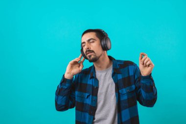 Dans eden ve şarkı söyleyen genç yakışıklı adam mavi bir arka planda kulaklıkla müzik dinliyor. Müzikten hoşlanan mutlu erkek.