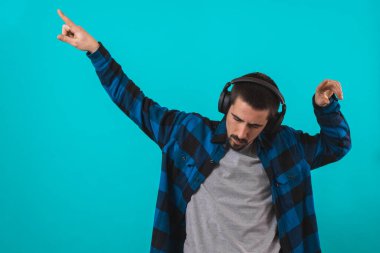 Dans eden ve şarkı söyleyen genç yakışıklı adam mavi bir arka planda kulaklıkla müzik dinliyor. Müzikten hoşlanan mutlu erkek.