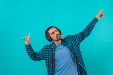 Dans eden ve şarkı söyleyen genç yakışıklı adam mavi bir arka planda kulaklıkla müzik dinliyor. Müzikten hoşlanan mutlu erkek.