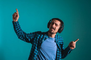 Dans eden ve şarkı söyleyen genç yakışıklı adam mavi bir arka planda kulaklıkla müzik dinliyor. Müzikten hoşlanan mutlu erkek.