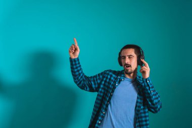 Dans eden ve şarkı söyleyen genç yakışıklı adam mavi bir arka planda kulaklıkla müzik dinliyor. Müzikten hoşlanan mutlu erkek.