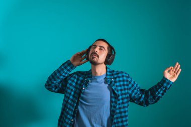 Dans eden ve şarkı söyleyen genç yakışıklı adam mavi bir arka planda kulaklıkla müzik dinliyor. Müzikten hoşlanan mutlu erkek.