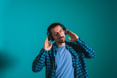 Dans eden ve şarkı söyleyen genç yakışıklı adam mavi bir arka planda kulaklıkla müzik dinliyor. Müzikten hoşlanan mutlu erkek.