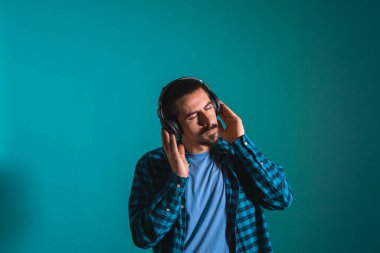 Dans eden ve şarkı söyleyen genç yakışıklı adam mavi bir arka planda kulaklıkla müzik dinliyor. Müzikten hoşlanan mutlu erkek.