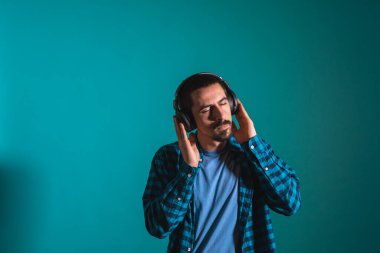 Dans eden ve şarkı söyleyen genç yakışıklı adam mavi bir arka planda kulaklıkla müzik dinliyor. Müzikten hoşlanan mutlu erkek.