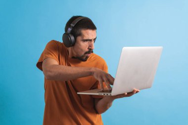 Oyun kulaklığı olan genç yakışıklı bir adam oyun oynayan dizüstü bilgisayarla mavi arka planda izole edilmiş..