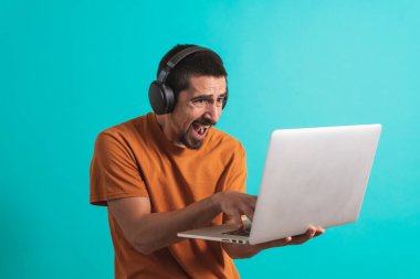 Oyun kulaklığı olan genç yakışıklı bir adam oyun oynayan dizüstü bilgisayarla mavi arka planda izole edilmiş..