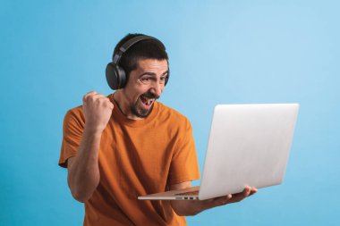 Oyun kulaklığı olan genç yakışıklı bir adam oyun oynayan dizüstü bilgisayarla mavi arka planda izole edilmiş..