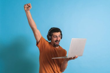 Oyun kulaklığı olan genç yakışıklı bir adam oyun oynayan dizüstü bilgisayarla mavi arka planda izole edilmiş..