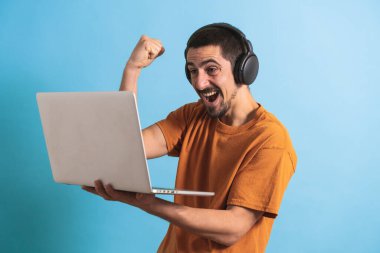 Oyun kulaklığı olan genç yakışıklı bir adam oyun oynayan dizüstü bilgisayarla mavi arka planda izole edilmiş..