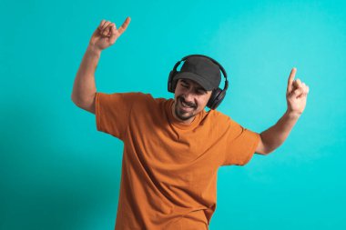 Dans eden ve şarkı söyleyen genç yakışıklı adam mavi bir arka planda kulaklıkla müzik dinliyor. Müzikten hoşlanan mutlu erkek.