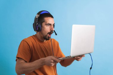 Oyun kulaklığı olan genç yakışıklı bir adam oyun oynayan dizüstü bilgisayarla mavi arka planda izole edilmiş..