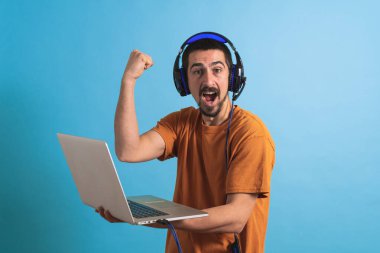Oyun kulaklığı olan genç yakışıklı bir adam oyun oynayan dizüstü bilgisayarla mavi arka planda izole edilmiş..