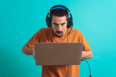 Oyun kulaklığı olan genç yakışıklı bir adam oyun oynayan dizüstü bilgisayarla mavi arka planda izole edilmiş..