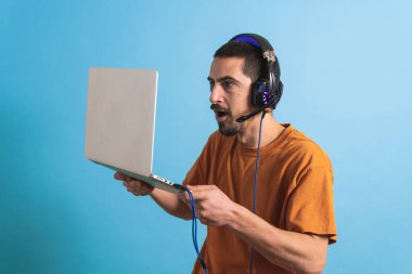 Oyun kulaklığı olan genç yakışıklı bir adam oyun oynayan dizüstü bilgisayarla mavi arka planda izole edilmiş..