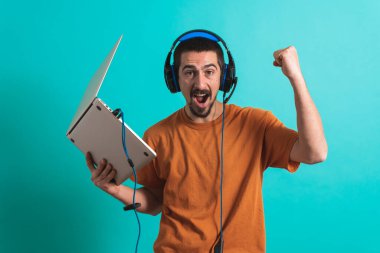 Oyun kulaklığı olan genç yakışıklı bir adam oyun oynayan dizüstü bilgisayarla mavi arka planda izole edilmiş..