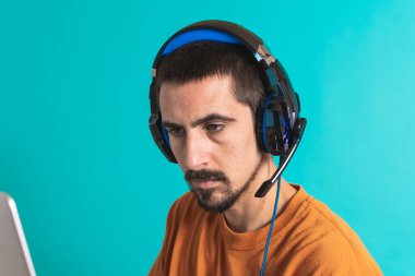 Oyun kulaklığı olan genç yakışıklı bir adam oyun oynayan dizüstü bilgisayarla mavi arka planda izole edilmiş..