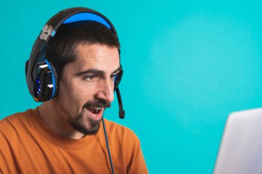 Oyun kulaklığı olan genç yakışıklı bir adam oyun oynayan dizüstü bilgisayarla mavi arka planda izole edilmiş..