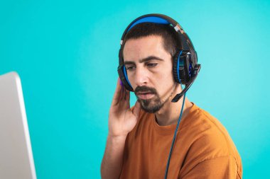Oyun kulaklığı olan genç yakışıklı bir adam oyun oynayan dizüstü bilgisayarla mavi arka planda izole edilmiş..
