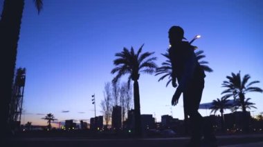 Genç havalı milenyum adamı, genç hippi kaykayıyla sahil şeridinde geziyor. Sinematografik aksiyon geniş çekim erkek kaykay numaraları yapmak Güneşli bir günde turistik bölgede.