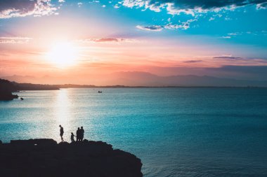 Ailenin silueti, sakin bir okyanusa yansıyan güneşin manzarasına bakıyor. Renkli gökyüzü gün doğumu.