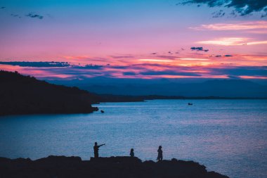 Ailenin silueti, sakin bir okyanusa yansıyan güneşin manzarasına bakıyor. Renkli gökyüzü gün doğumu.