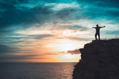 İnsan silueti uçurumun kenarında duruyor. Güzel renkli gökyüzü ile denize bakıyor..