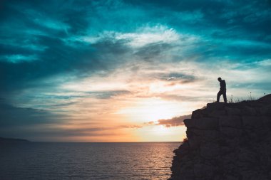 İnsan silueti uçurumun kenarında duruyor. Güzel renkli gökyüzü ile denize bakıyor..
