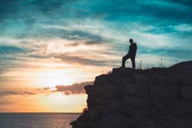 İnsan silueti uçurumun kenarında duruyor. Güzel renkli gökyüzü ile denize bakıyor..