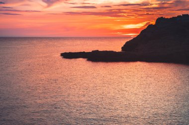 Sahil kayalıklarının ve denizin üzerinde turuncu renk yansımasıyla gün doğumu. Gün batımı ve renkli gökyüzü ile ufka karşı körfezin çarpıcı panoramik görüntüsü