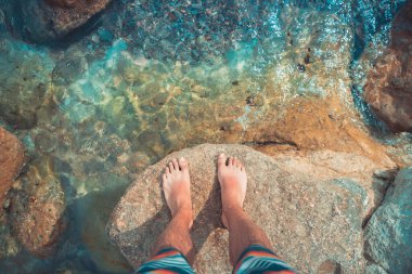Çıplak ayaklı genç adamın bakış açısı deniz sahilindeki turkuaz suda duruyor. Akdeniz yaz tatillerinde dinlenir.