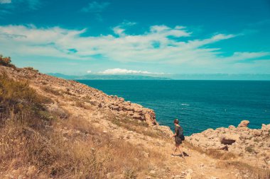 Güzel bir kadın, dağların üzerinde ve derin mavi okyanusun üzerinde inanılmaz renkli bir gökyüzünün kenarında yürüyor. Gün batımında deniz manzarası.